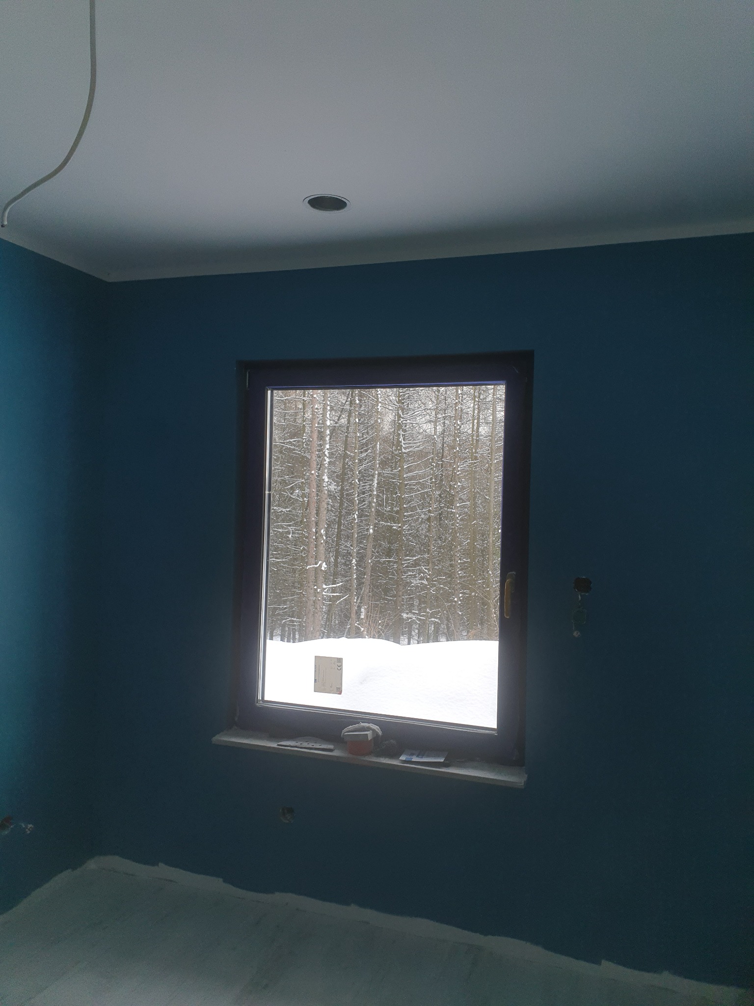 Pomieszczenie po malowaniu ścian na kolor morski, widoczne niedociągnięcia przy podłodze, okno z widokiem na zimowy las, sufit biały z punktowym oświetleniem.