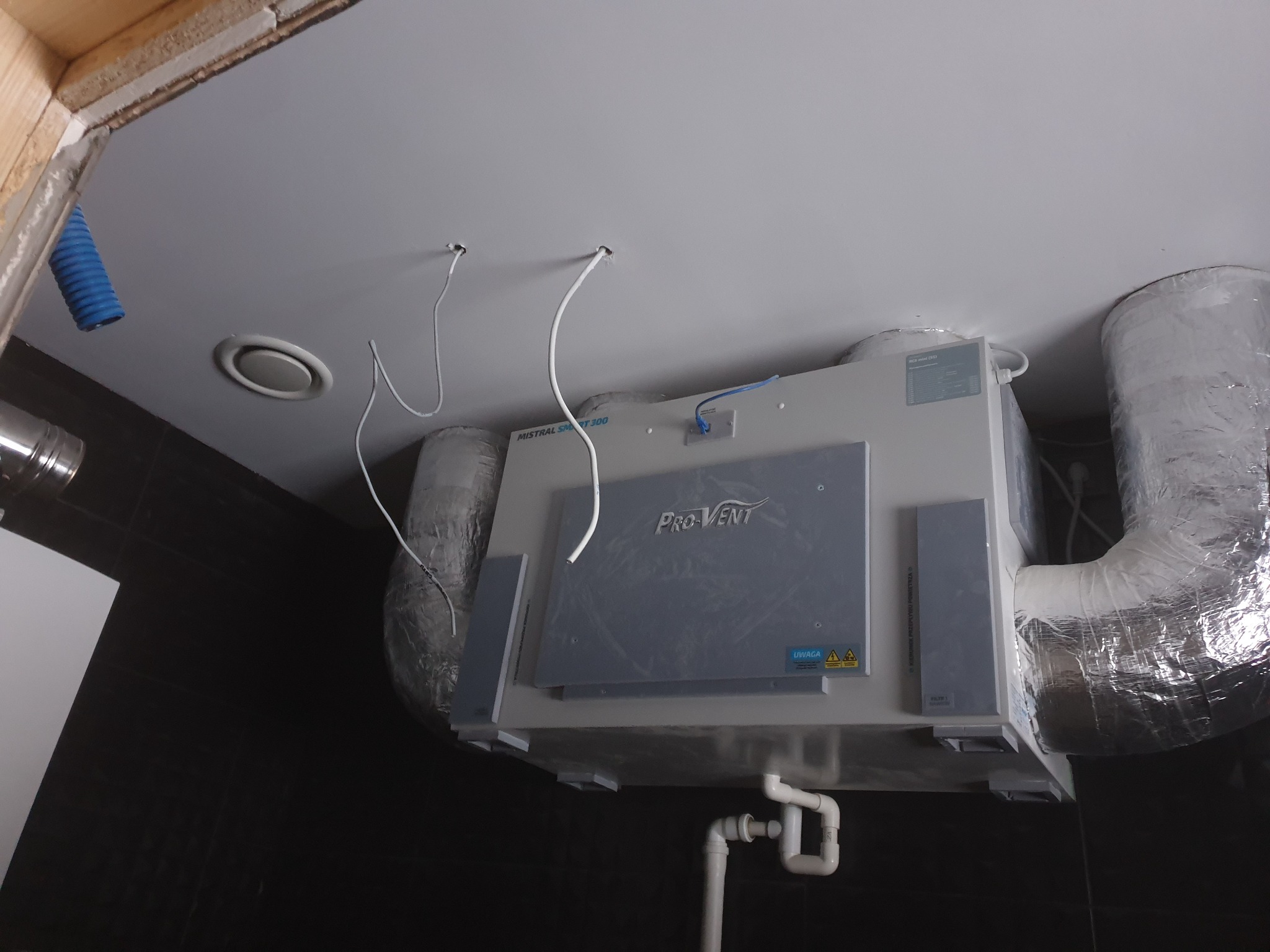 Zamontowana centrala wentylacyjna Pro-Vent Mistral Smart 300 na suficie podwieszanym, widoczne przewody wentylacyjne w izolacji aluminiowej, rury odprowadzające skropliny oraz wystające kable...