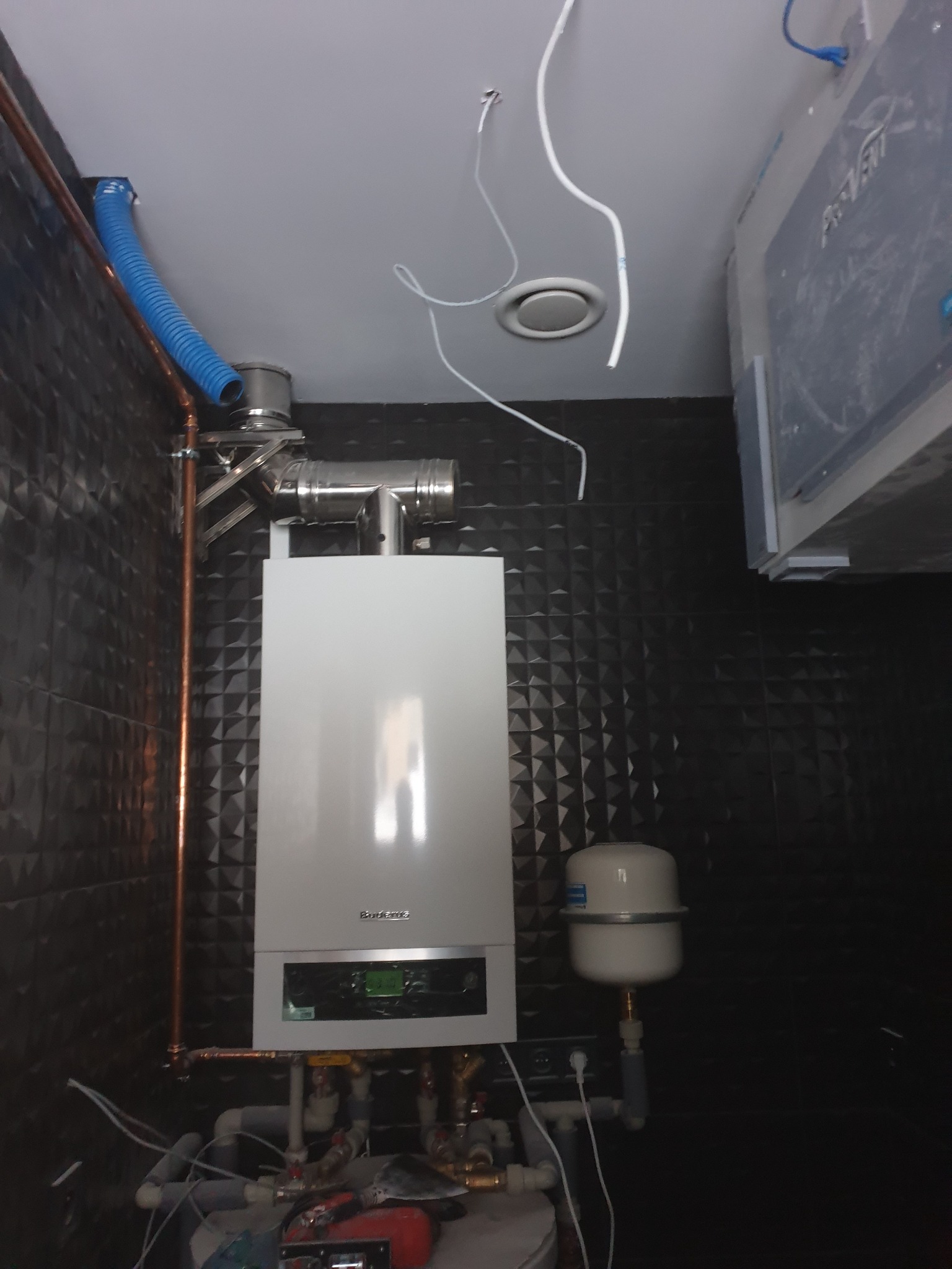Zamontowany, biały piec gazowy marki Buderus z podłączeniami miedzianymi i niebieskim przewodem wentylacyjnym na tle ściany z czarnych, trójwymiarowych płytek.