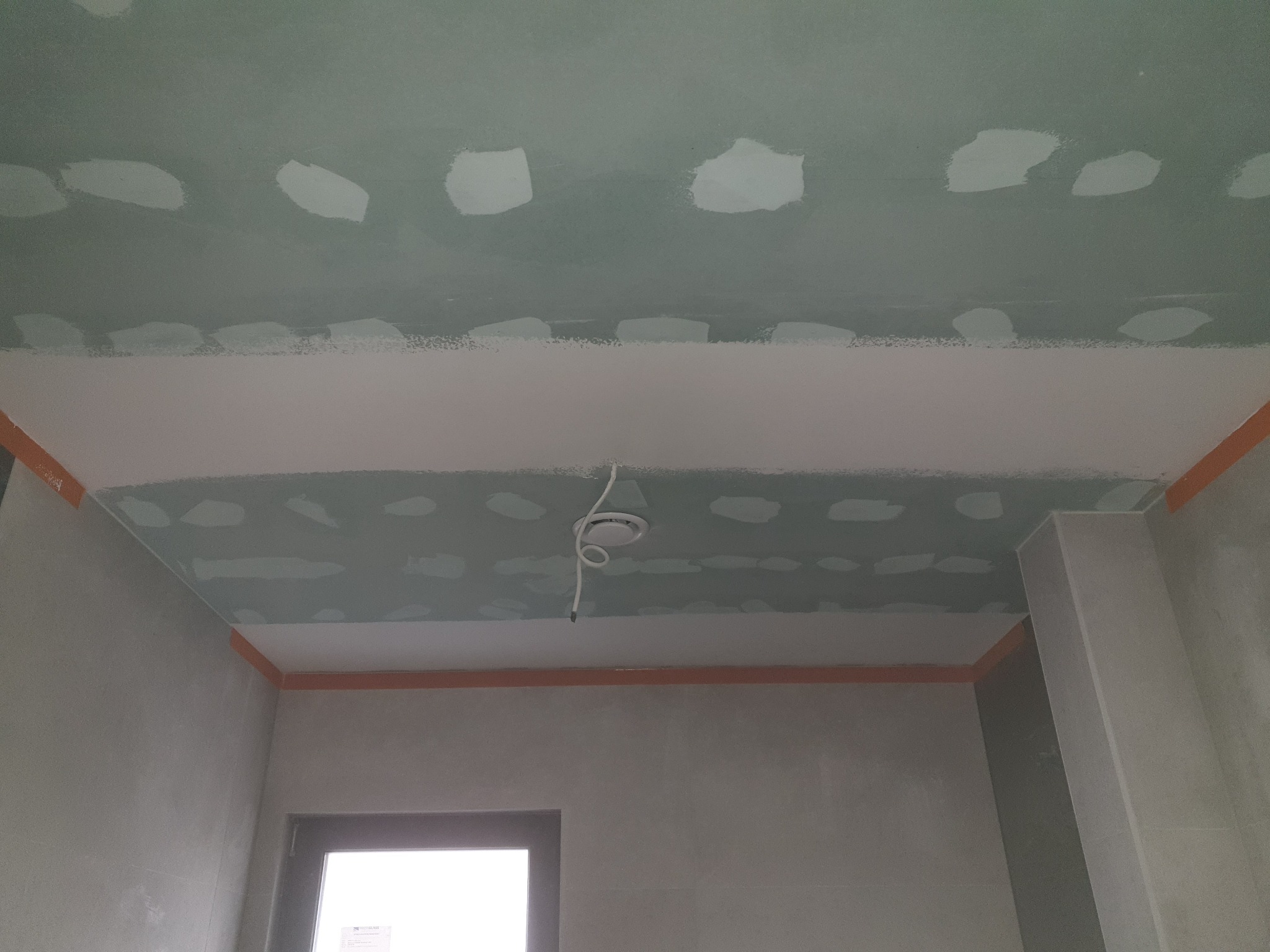 Sufit z płyt gipsowo-kartonowych w trakcie wykańczania, widoczne łączenia i miejsca po wkrętach wypełnione masą szpachlową, zabezpieczone taśmą malarską narożniki.