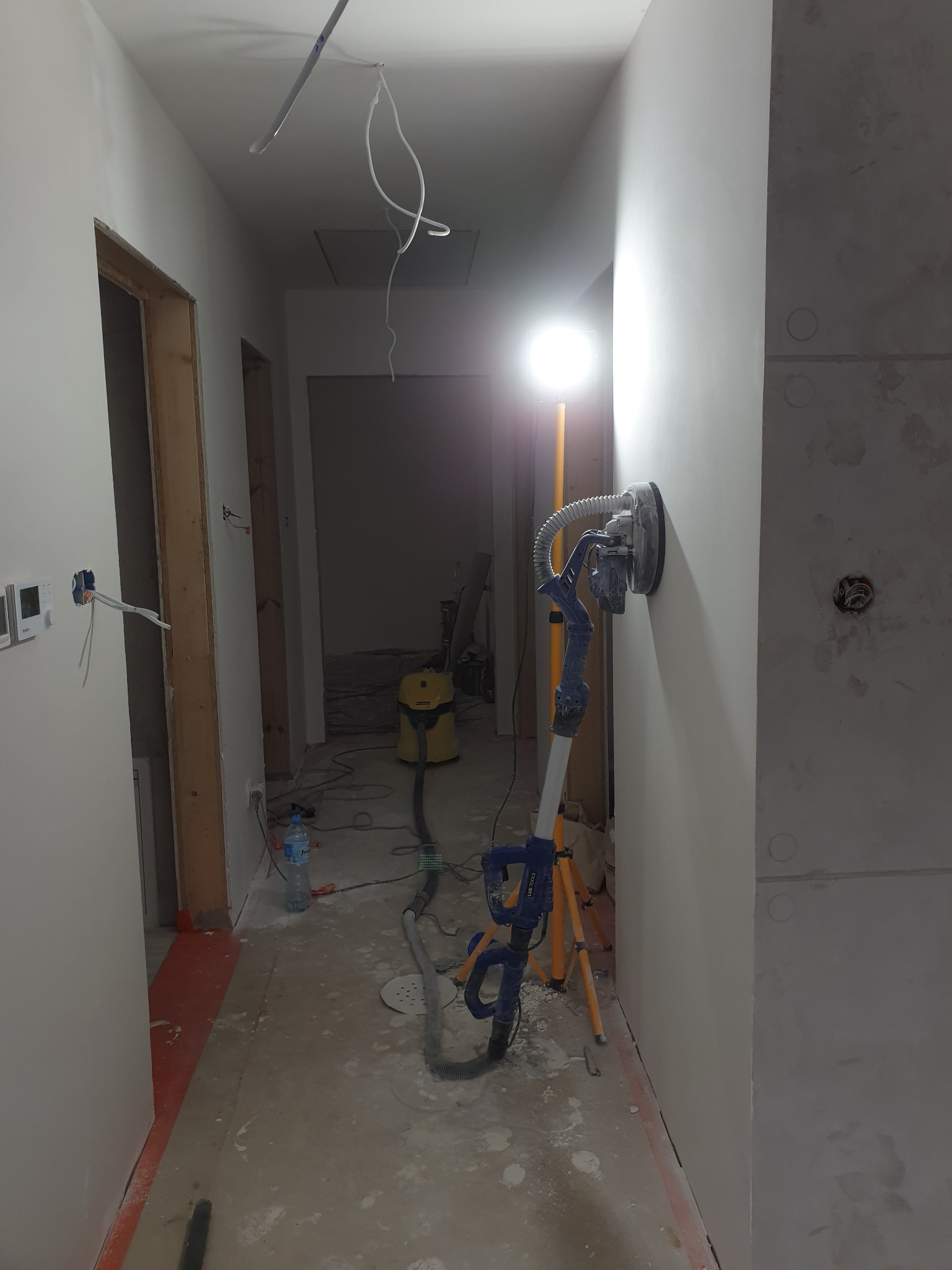 Korytarz w trakcie prac wykończeniowych, widoczna szlifierka do gipsu oparta o ścianę, żółty odkurzacz przemysłowy, przewody elektryczne zwisające z sufitu, surowy stan wykończenia.