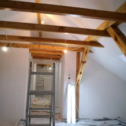 Poddasze w trakcie remontu, widoczne drewniane belki konstrukcyjne, świeżo pomalowane białe ściany, rozłożona folia ochronna na podłodze, drabina aluminiowa i lampa budowlana.