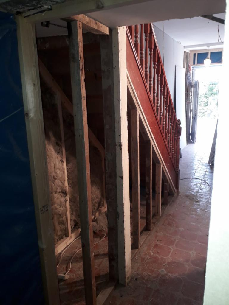 Klatka schodowa w trakcie remontu: widoczne drewniane elementy konstrukcyjne, izolacja, czerwona balustrada oraz stare, ceglane płytki podłogowe.