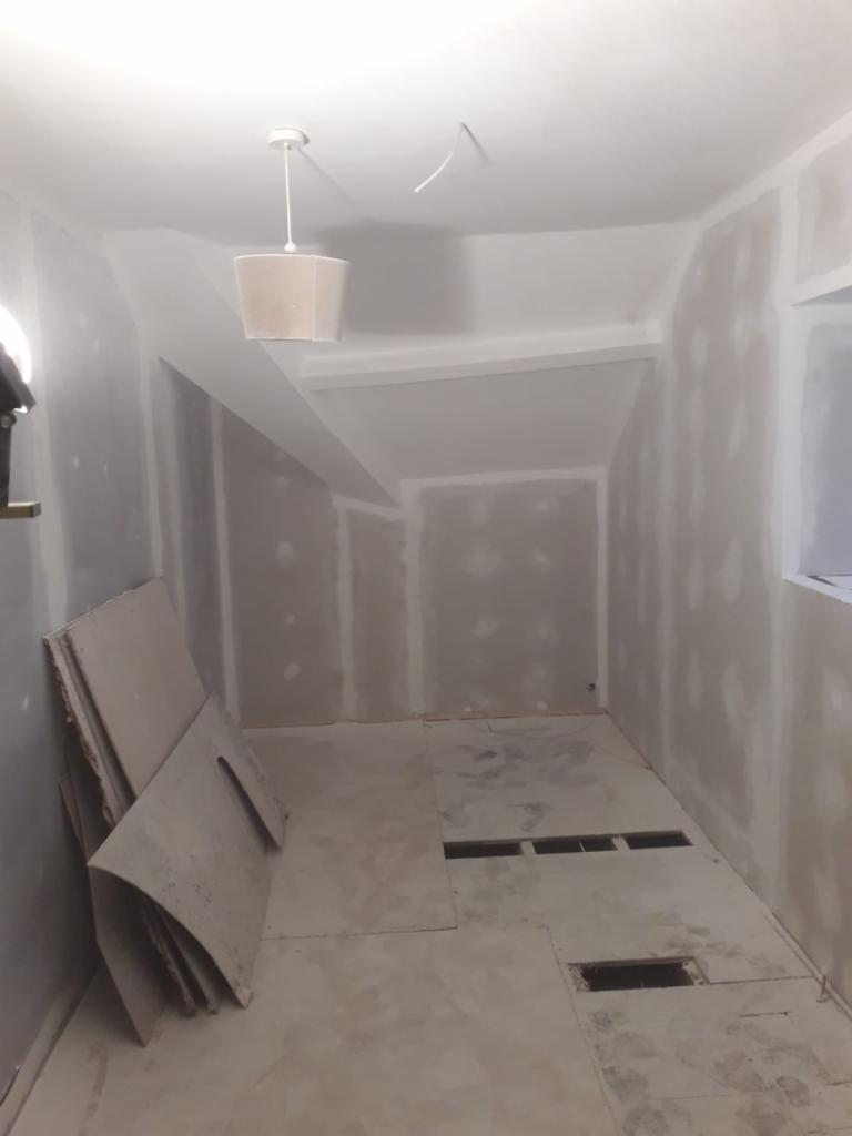 Pokój w trakcie remontu: ściany po szpachlowaniu, przygotowane do malowania, na podłodze płyty, widoczne otwory w podłodze, lampa sufitowa.