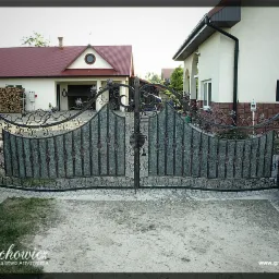 Dwuskrzydłowa brama wjazdowa z elementami kutymi i wypełnieniem imitującym kamień, osadzona na słupkach z dekoracyjnymi kulami, na tle dwóch budynków mieszkalnych.