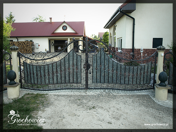 Dwuskrzydłowa brama wjazdowa z elementami kutymi i wypełnieniem imitującym kamień, osadzona na słupkach z dekoracyjnymi kulami, na tle dwóch budynków mieszkalnych.