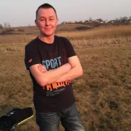 Mężczyzna w koszulce sportowej, z założonymi rękami, na tle pola i zabudowań w oddali, w promieniach zachodzącego słońca.