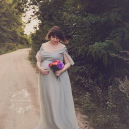 MWFotografia - Kobieta w zwiewnej, błękitnej sukni ciążowej, trzymająca bukiet kolorowych kwiatów, spaceruje po polnej drodze w otoczeniu zieleni.