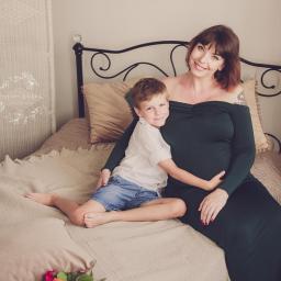 MWFotografia - Uśmiechnięta kobieta w ciemnozielonej sukni ciążowej siedzi na łóżku, obejmowana przez syna w białej koszuli i dżinsowych spodenkach, w tle widoczny parawan i roślina doniczkowa.