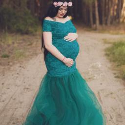 MWFotografia - Kobieta w ciąży w szmaragdowej sukni z koronki i tiulu, z wiankiem z róż na głowie, pozuje na leśnej ścieżce.