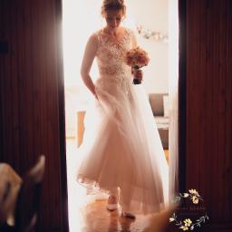 MWFotografia - Panna młoda w koronkowej sukni wchodzi przez drewniane drzwi, trzymając bukiet różowych i białych róż.