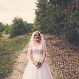 MWFotografia - Panna młoda w białej sukni z koronkowym gorsetem i welonem, trzyma bukiet różowych i kremowych róż na leśnej ścieżce.