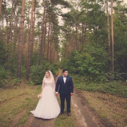 MWFotografia - Para młoda trzymająca się za ręce na leśnej ścieżce, panna młoda w koronkowej sukni i welonie, pan młody w granatowym garniturze z muchą, wysokie sosny w tle, stonowana kolorystyka.