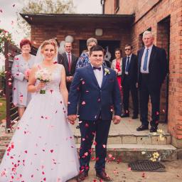 MWFotografia - Uśmiechnięta para młoda trzymająca się za ręce, obsypywana konfetti w kształcie serc, na tle ceglanego budynku i gości weselnych.