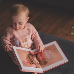 MWFotografia - Małe dziecko w różowym swetrze z aplikacjami zwierząt ogląda album ze zdjęciem noworodka w czepku, leżącego na kocyku.