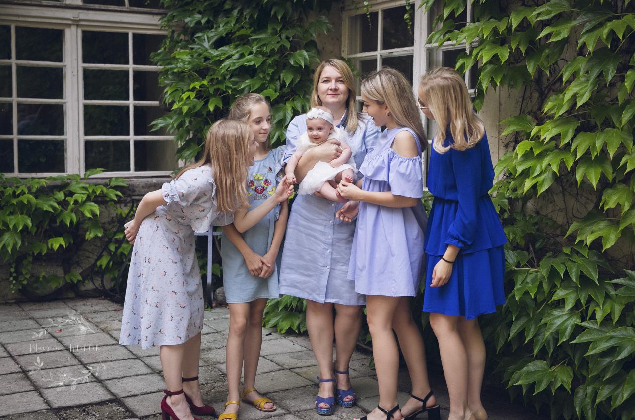 Kobieta w niebieskiej sukience trzyma niemowlę w otoczeniu czterech dziewcząt w różnych odcieniach niebieskiego, na tle ściany porośniętej bluszczem.