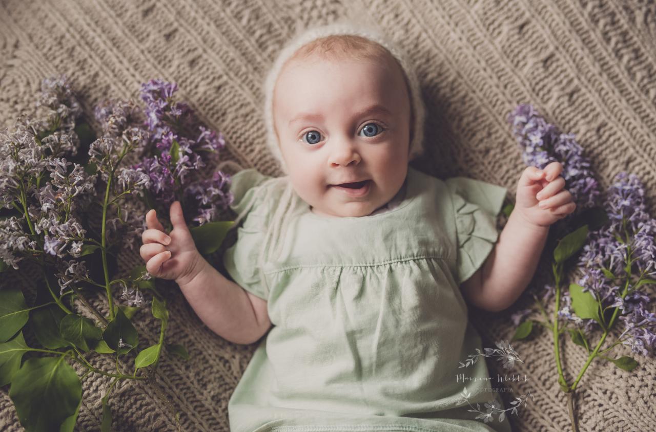 Niemowlę z dużymi niebieskimi oczami leżące na beżowym kocu w warkocze, ubrane w zieloną sukienkę i białą czapeczkę, otoczone gałązkami bzu.