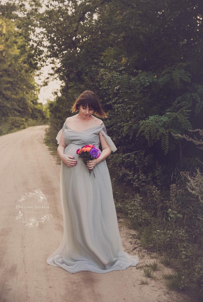 Kobieta w zwiewnej, błękitnej sukni ciążowej, trzymająca bukiet kolorowych kwiatów, spaceruje po polnej drodze w otoczeniu zieleni.