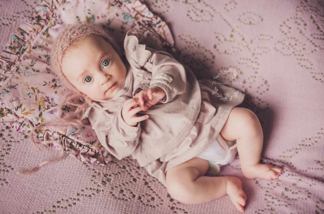 Niemowlę z jasnymi oczami leży na plecach na różowym, wzorzystym kocyku, ubrane w beżową sukienkę i czapeczkę, z delikatnie splecionymi dłońmi.