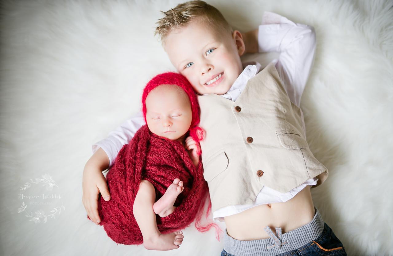 Noworodek zawinięty w czerwoną dzianinę leży obok uśmiechniętego starszego brata na białym futrzanym tle.