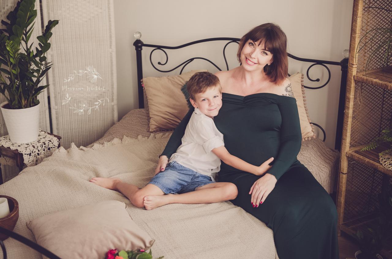 Uśmiechnięta kobieta w ciemnozielonej sukni ciążowej siedzi na łóżku, obejmowana przez syna w białej koszuli i dżinsowych spodenkach, w tle widoczny parawan i roślina doniczkowa.