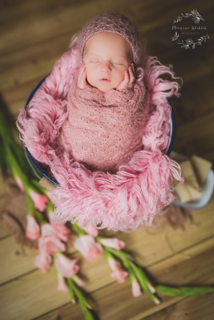 Noworodek owinięty w różowy koc, śpiący w metalowej misce z kwiatami. Fotografia z góry, ciepłe oświetlenie, drewniane tło.