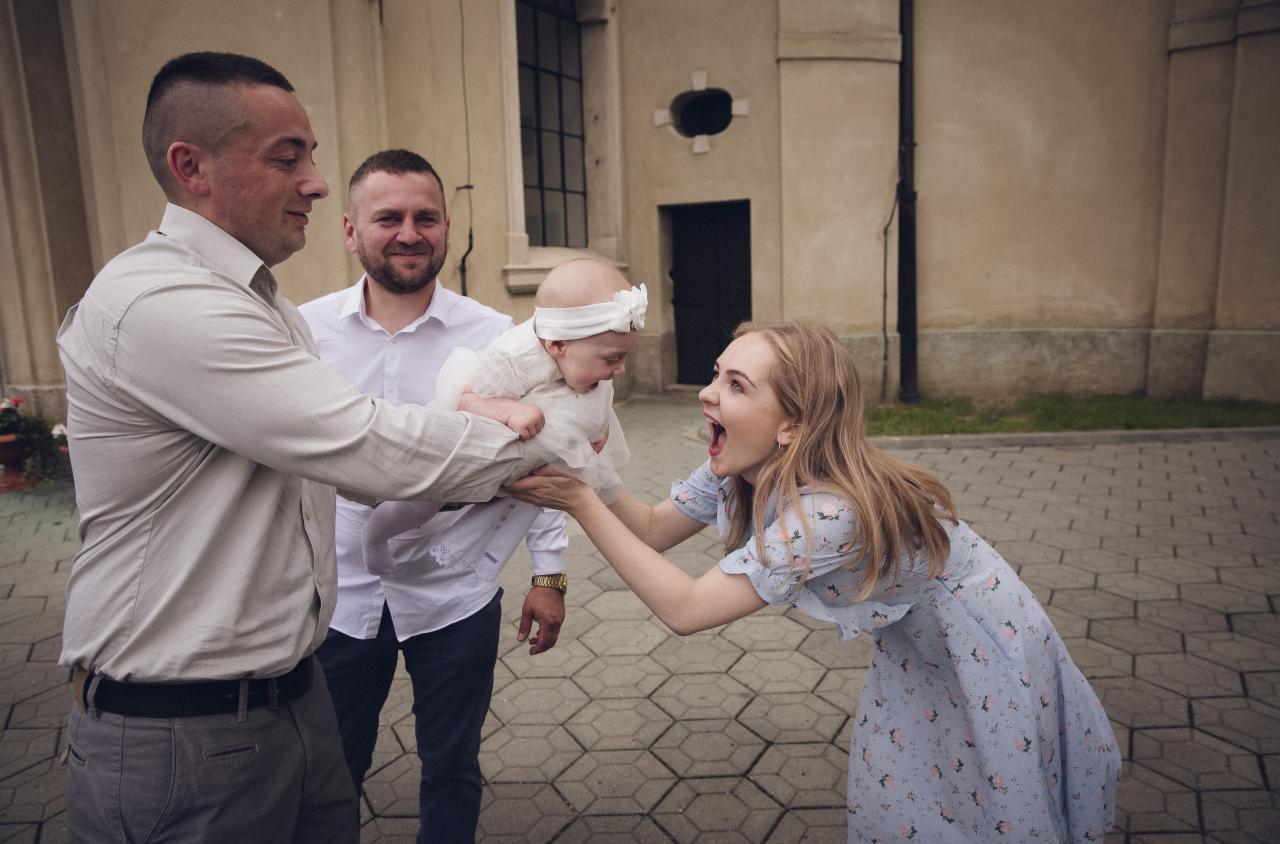Rodzinne ujęcie z uroczystości: mężczyzna trzyma niemowlę w białej sukience, a kobieta z entuzjazmem wyciąga ręce, obok stoi uśmiechnięty mężczyzna, na tle kościoła.