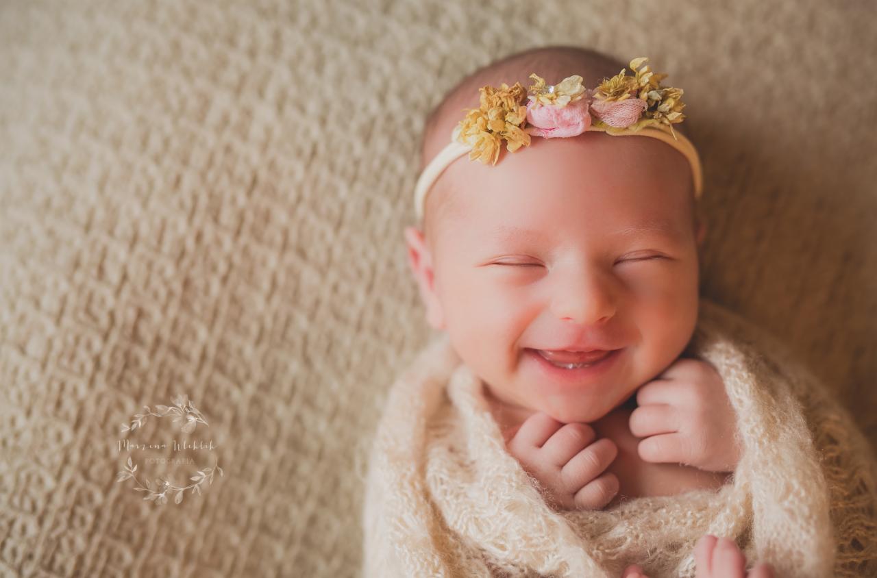 Uśmiechnięty noworodek, owinięty w beżowy ażurowy kocyk, z zamkniętymi oczami i kwiatową opaską na głowie, leży na teksturowanym tle.