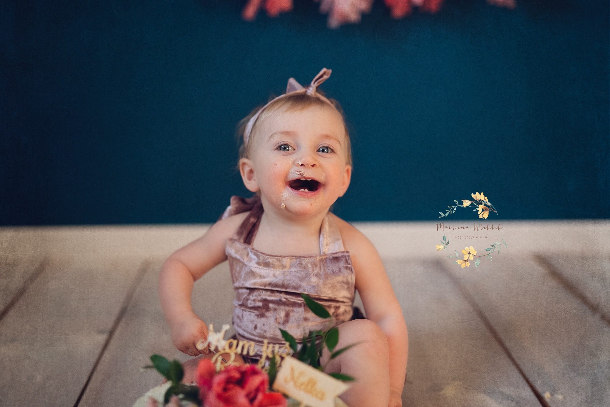 Uśmiechnięta roczna dziewczynka w welurowym ubranku, z resztkami tortu na twarzy, siedzi na drewnianej podłodze przed tortem z kwiatami i napisem 'Nelka', na tle ciemnoniebieskiej ściany.