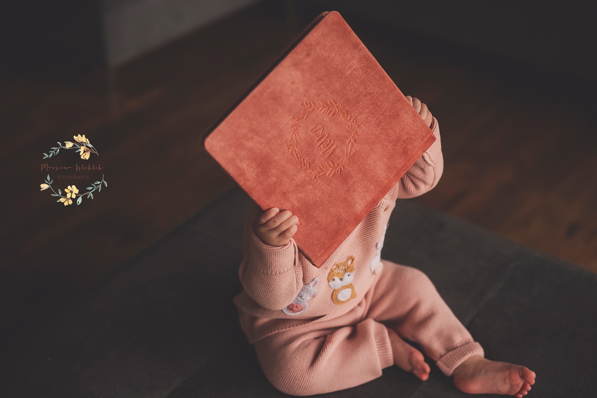 Niemowlę w różowym ubranku siedzi, trzymając przed twarzą kwadratowy album w kolorze łososiowym z wytłoczonym napisem otoczonym wiankiem.