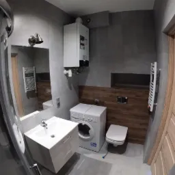 Nowoczesna łazienka w szarościach z drewnianymi akcentami, umywalka z szafką, pralka i podwieszana toaleta, widoczny piec gazowy na ścianie.