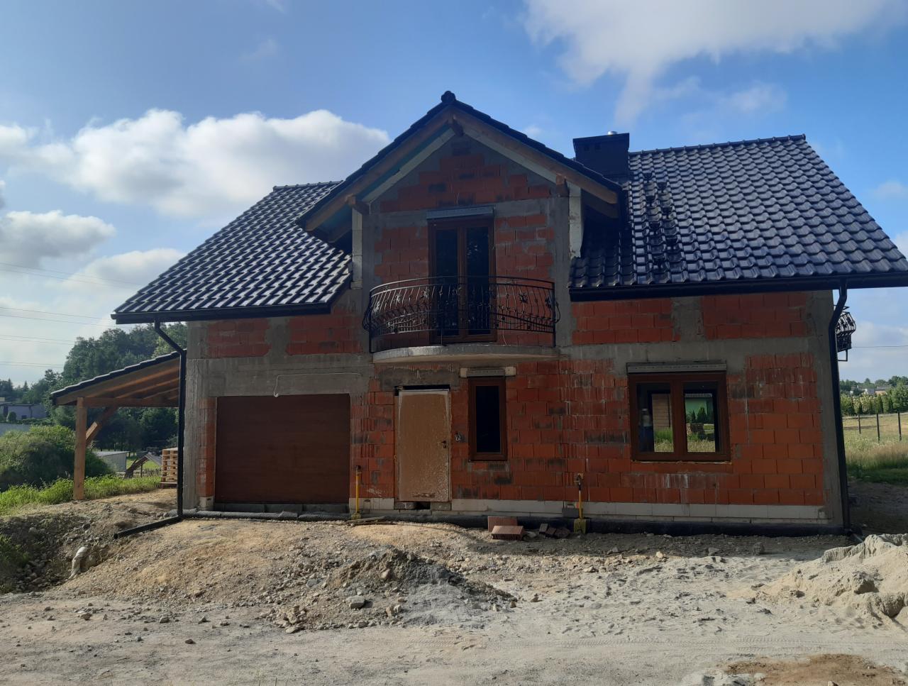 Niedokończony dom jednorodzinny z czerwonej cegły z czarnym dachem z dachówki ceramicznej, balkonem z ozdobną balustradą i drewnianą bramą garażową, widoczny w słoneczny dzień na tle błękitnego nieba.