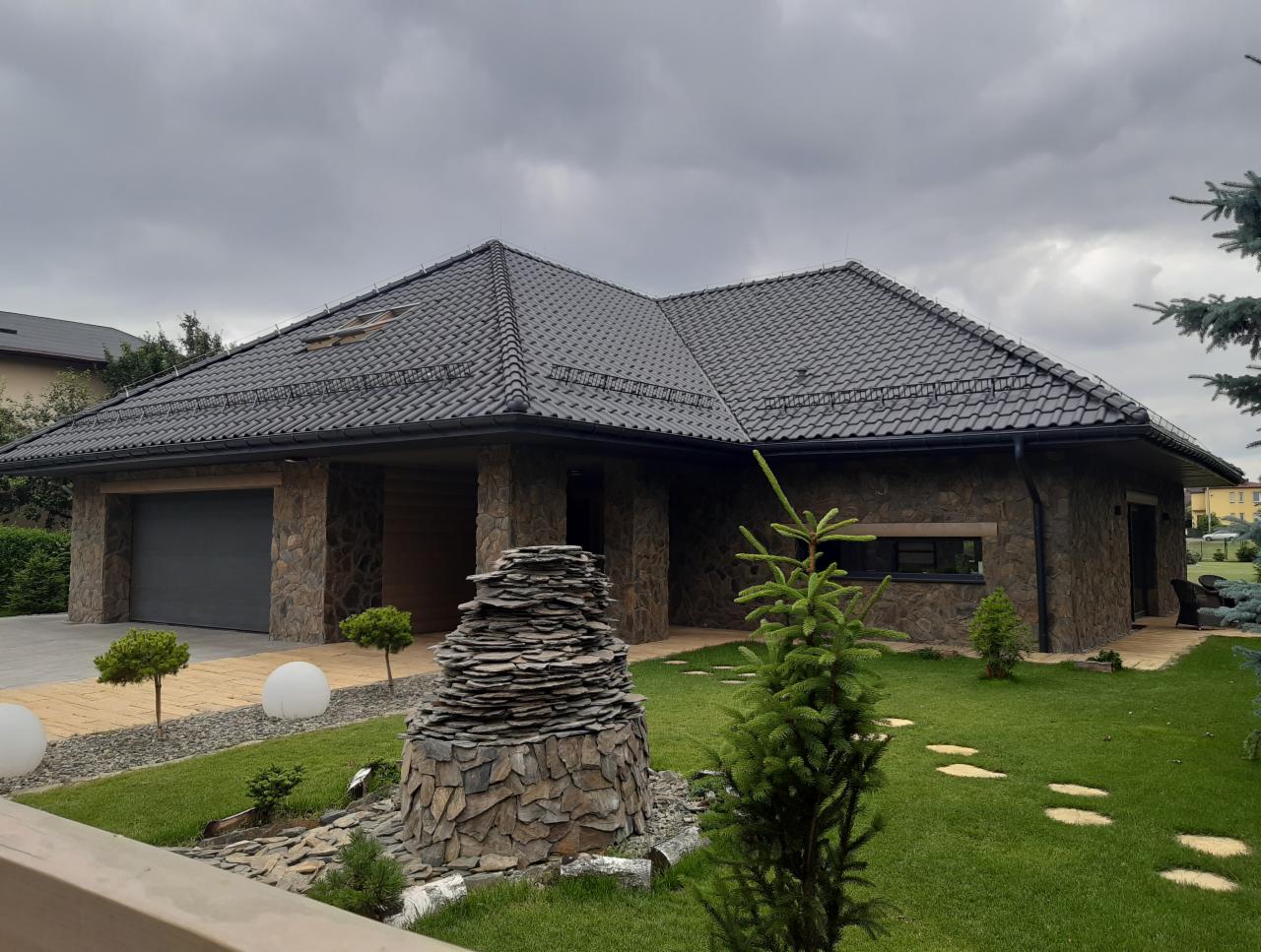 Nowoczesny dom jednorodzinny z kamienną elewacją i ciemną dachówką, otoczony zadbanym trawnikiem z dekoracyjnymi kamieniami i oświetleniem.
