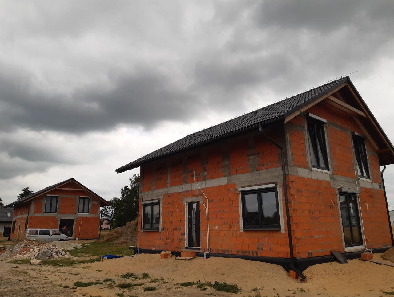 Dwa domy w stanie surowym otwartym, z cegły ceramicznej, z czarną dachówką, okna wstawione, teren wokół przygotowany, pochmurne niebo w tle.