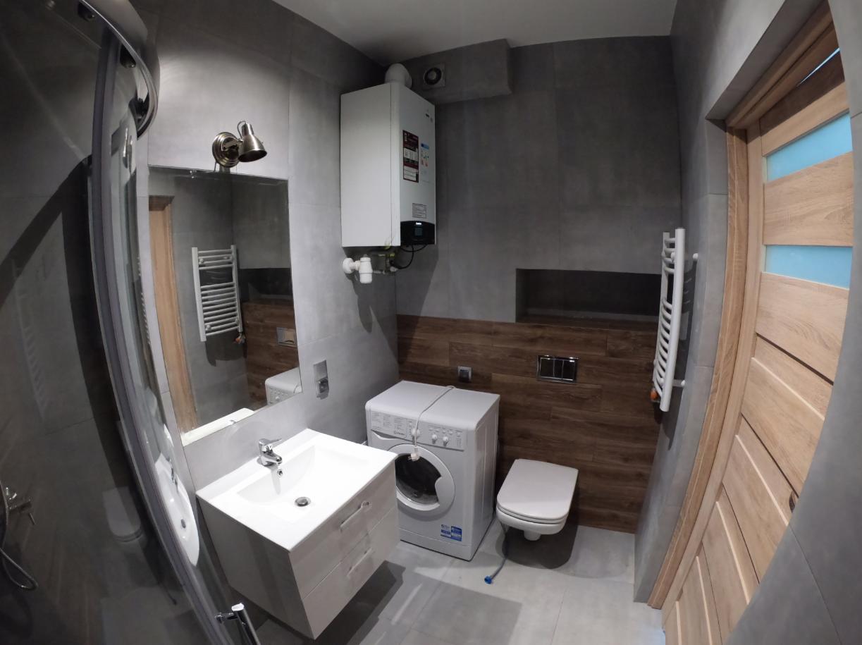 Nowoczesna łazienka w szarościach z drewnianymi akcentami, umywalka z szafką, pralka i podwieszana toaleta, widoczny piec gazowy na ścianie.