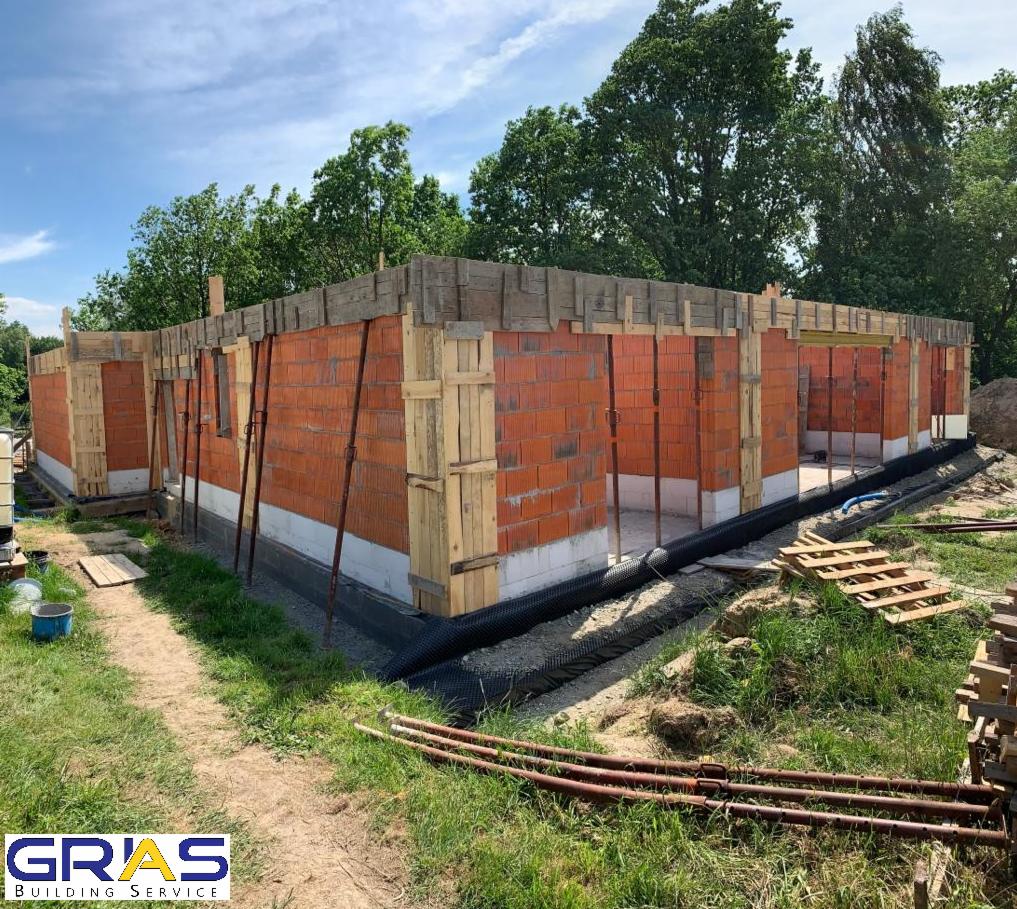 Wznoszenie ścian parterowego domu z czerwonej cegły, widoczne drewniane szalunki i podpory, fundament zabezpieczony folią, w tle zieleń drzew i błękitne niebo.