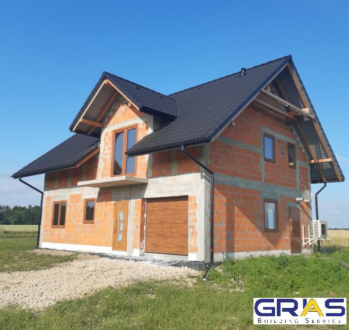 Niedokończony dom murowany z czarnym dachem i brązową bramą garażową, widoczne pustaki i niezabezpieczona elewacja, teren wokół domu częściowo zagospodarowany.