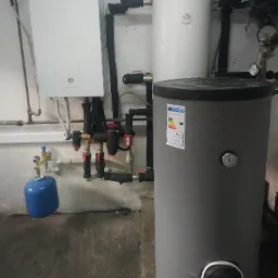 Instalacja z białym bojlerem Panasonic, szarym zbiornikiem z etykietą energetyczną i niebieską butlą, widoczne liczne rury i zawory w pomieszczeniu technicznym.