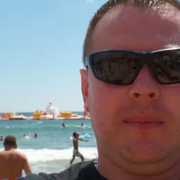 Mężczyzna w okularach przeciwsłonecznych robi selfie na plaży z dmuchanym parkiem wodnym w tle, błękitne niebo i woda.
