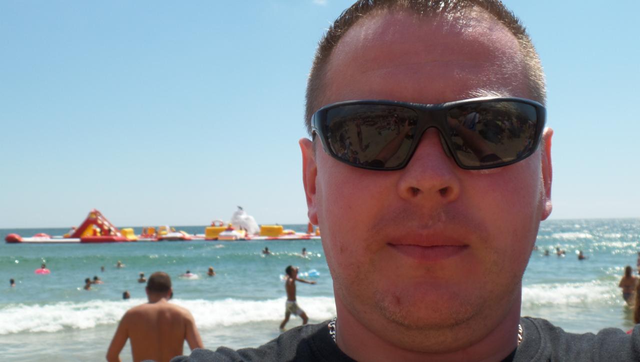 Mężczyzna w okularach przeciwsłonecznych robi selfie na plaży z dmuchanym parkiem wodnym w tle, błękitne niebo i woda.