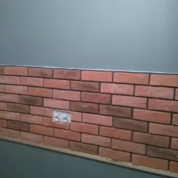 Ściana z imitacji cegły w odcieniach czerwieni i brązu, częściowo położona, z widocznym gniazdkiem elektrycznym i szarą ścianą powyżej.