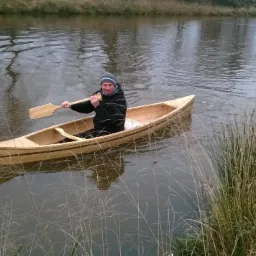 Mężczyzna płynie drewnianą łodzią wiosłową po rzece, otoczony roślinnością brzegową. Łódź z jasnego drewna z ciemniejszymi paskami. Wiosło w dłoni.