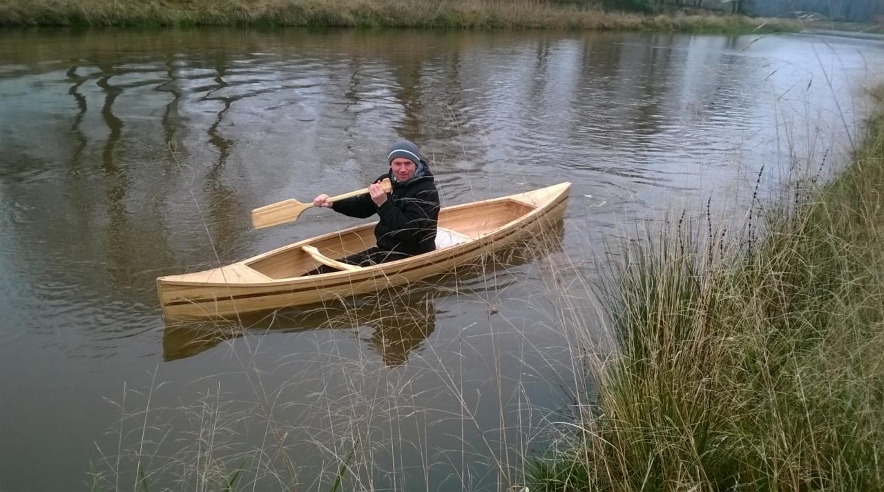 Mężczyzna płynie drewnianą łodzią wiosłową po rzece, otoczony roślinnością brzegową. Łódź z jasnego drewna z ciemniejszymi paskami. Wiosło w dłoni.