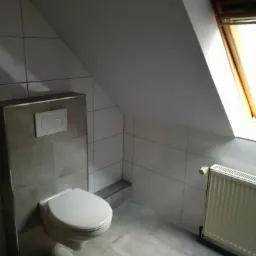 Wykończona łazienka na poddaszu z wiszącą toaletą, szarymi płytkami podłogowymi i ściennymi oraz grzejnikiem pod oknem dachowym.