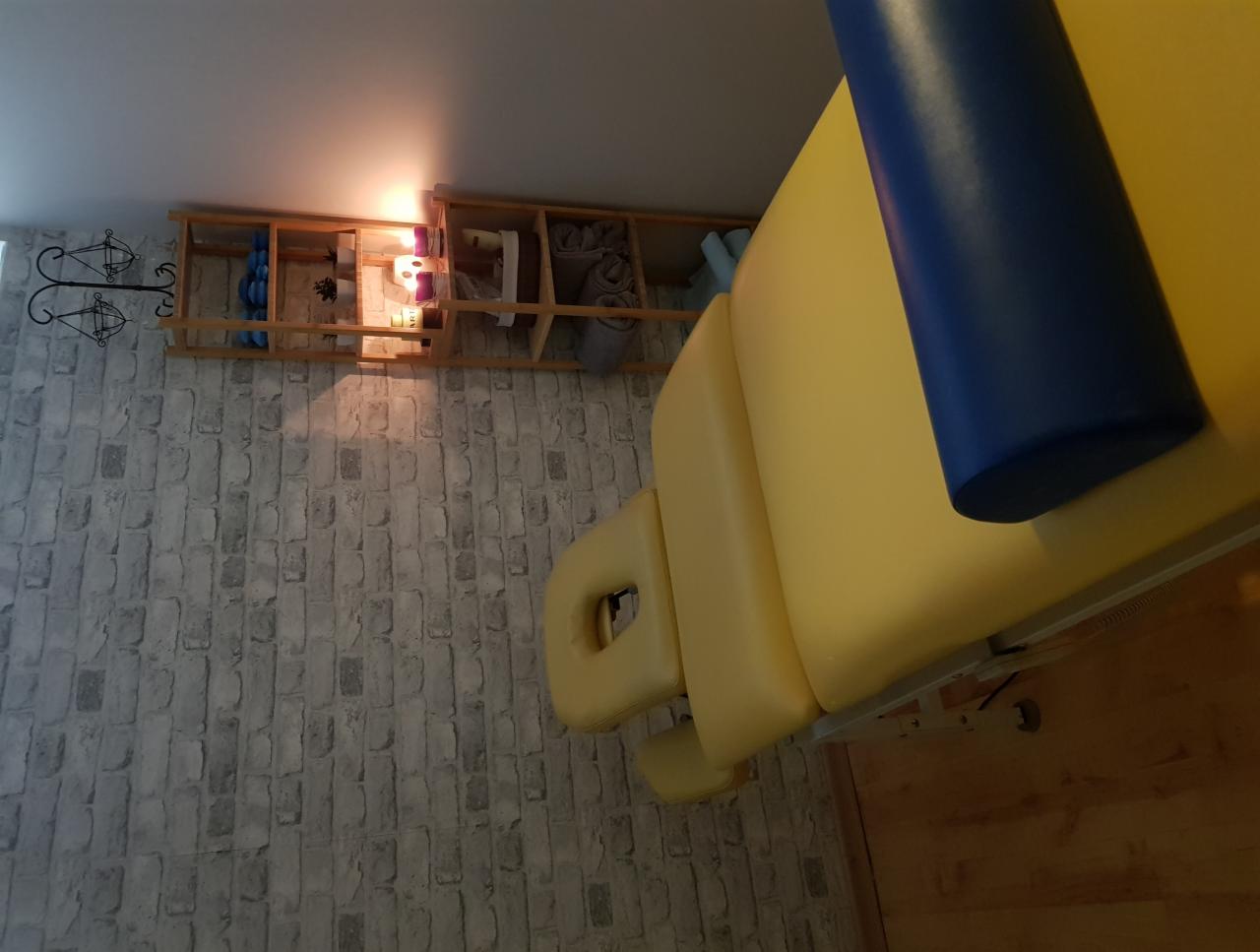 Żółty stół do zabiegów z niebieskim wałkiem, widok z boku, na ścianie półka z akcesoriami i dekoracjami.