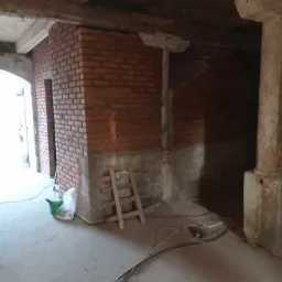 Wnętrze w trakcie remontu z surową ceglaną ścianą, drewnianymi belkami stropowymi i betonową podłogą, widoczny mały drewniany drabina oparta o ścianę.
