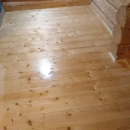 Świeżo polakierowana drewniana podłoga w domu z bali, z widocznym wałkiem malarskim w rogu.