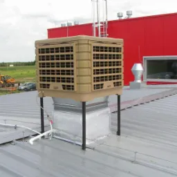 Beżowy klimatyzator przemysłowy na dachu z blachy trapezowej, widoczne przewody i elementy montażowe, w tle czerwony budynek i sprzęt budowlany.