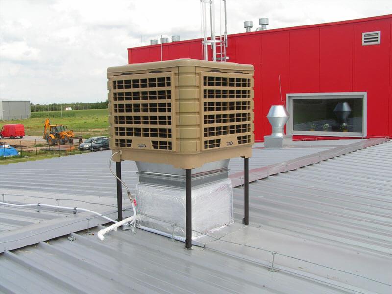 Beżowy klimatyzator przemysłowy na dachu z blachy trapezowej, widoczne przewody i elementy montażowe, w tle czerwony budynek i sprzęt budowlany.