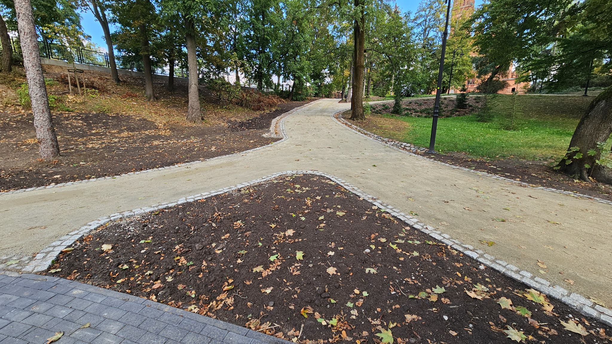 Rozwidlenie nowo utwardzonych ścieżek parkowych, obrzeża z kostki granitowej, w tle drzewa i budynek z czerwonej cegły.